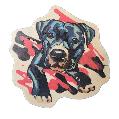 Pancake art of a black puppy named Hilde, shown lying down with front paws extended forward, head up and facing the viewer, with floppy ears and a tan muzzle; the dog appears to be a large-breed puppy based on its proportions and facial structure. Rendered by Dan with fine dark outlines and layered…