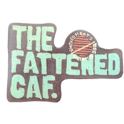 Pancake art depicting the text 'THE FATTENED CAF.' in large, bold teal block letters on a dark brown background, with a circular logo element in the upper right showing a stylized round grill grate in red and brown with two crossed drumsticks or skewers, surrounded by the curved text 'SMOKED MEATS'…
