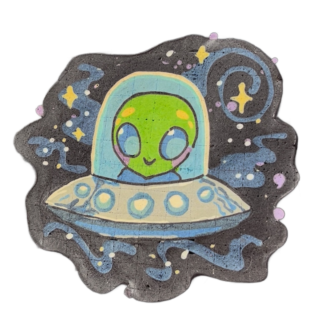 Pancake art depicting Trav, a green-skinned alien character with large blue eyes, orange cheek markings, pink cheeks, and a lavender scarf, seated inside a domed flying saucer with circular porthole details along its rim, set against a dark background with blue swirling lines, scattered white and y…