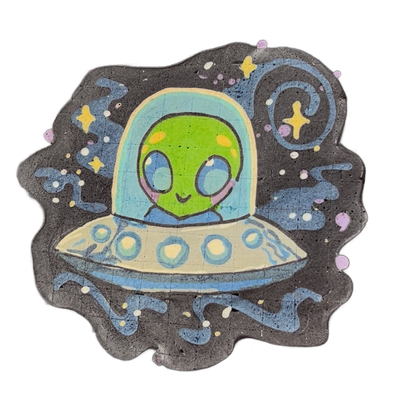Pancake art depicting Trav, a green-skinned alien character with large blue eyes, orange cheek markings, pink cheeks, and a lavender scarf, seated inside a domed flying saucer with circular porthole details along its rim, set against a dark background with blue swirling lines, scattered white and y…