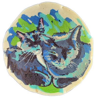 Pancake art depicting two cats curled closely together, each facing forward with visible whiskers, ears, and yellow-green eyes, set against a background of overlapping brush-stroke shapes. The piece uses bold dark outlines filled with blue, indigo, teal, and green batter, with lighter cream and whi…