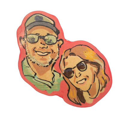 Pancake art depicting two adult human faces side by side, both wearing sunglasses; the figure on the left presents with darker skin tone, a beard, a pearl or beaded necklace, a baseball cap, and a green shirt, while the figure on the right has lighter skin tone, shoulder-length wavy hair with warm…