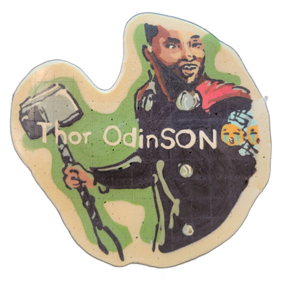 Pancake art depicting Thor Odinson: a bearded man with short dark hair, wearing dark armor with a red cape, holding a large hammer (Mjolnir) by a twisted handle in his left hand, with the text 'Thor OdinSON' written across the center and two small crying-face emoji-style icons to the right of the t…