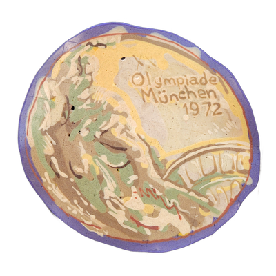 Pancake art depicting what appears to be a replica of an Olympic gold medal, showing a classical figure in profile on the left side and the text 'Olympiade München 1972' on the right, referencing the 1972 Munich Summer Olympics medal design. The piece uses bold outlines and broad fill areas in yell…