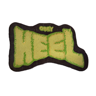 Pancake art depicting the word 'HEEL' in large block letters, with 'OBEY' written in smaller rounded letters above it, all rendered in a graffiti-style layout. The lettering is filled in yellow-tan batter with bold green outlines, set against a dark brown background border; the surface shows fine g…