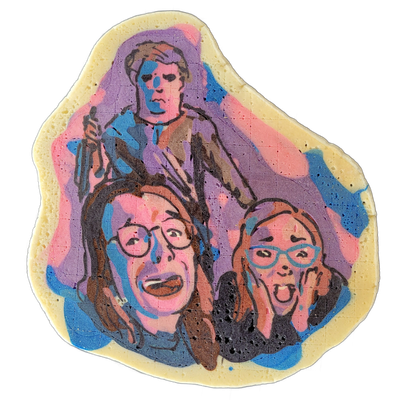 Pancake art titled 'MICHAEL!' depicting three figures: a taller figure in the background wearing a hooded or draped garment and holding an object in one hand, and two figures in the foreground — one on the left with long dark hair, round glasses, and an open-mouthed expression, and one on the right…