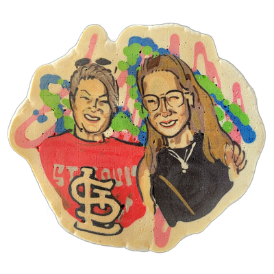 Pancake art depicting two people posed side by side and smiling: on the left, a person with short dark hair wearing a red shirt printed with 'St. Louis' text and the St. Louis Cardinals interlocking 'StL' logo; on the right, a person with long straight light-brown hair and round glasses wearing a d…