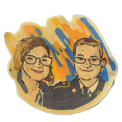 Pancake art depicting two people shown from the shoulders up, side by side: on the left, a person with shoulder-length wavy hair and rectangular glasses; on the right, a person with short hair and rectangular glasses; both wear dark gray clothing and appear to be smiling slightly, with small gold o…