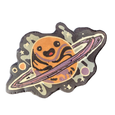 Pancake art depicting Saturnina, a JoP original character rendered as a cartoon planet with Saturn-style rings and a round face featuring two dot eyes, a small open mouth with a red accent, and dark swirling stripe markings across an orange body. The piece uses bold black outlines, a palette of ora…