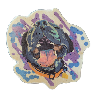 Pancake art depicting a dog's face, identified as Milo, centered within what appears to be a round helmet or visor shape rendered in dark batter, with the dog's face showing a pink-toned muzzle, dark fur markings, and teal-highlighted ears or fur at the top. The piece is drawn with bold dark outlin…