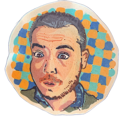 Pancake art of a face labeled 'Catastrophe' in the artist's canon, depicting a man with short dark hair, a mustache, and a short beard, wearing what appears to be a blue jacket, with wide-open eyes and a slightly open mouth giving him a startled expression, set against a checkered background of blu…