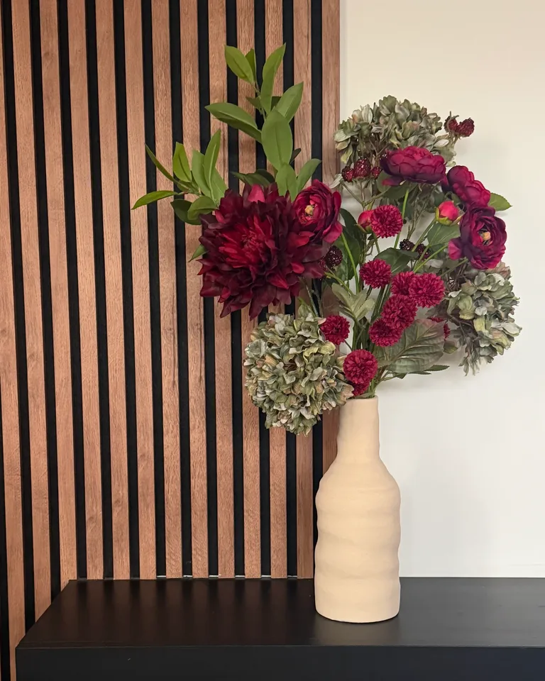 Zijden boeket Rubina met burgundy bloemen en groene hortensia in een beige vaas op een zwarte wandkast naast een houten lattenwand