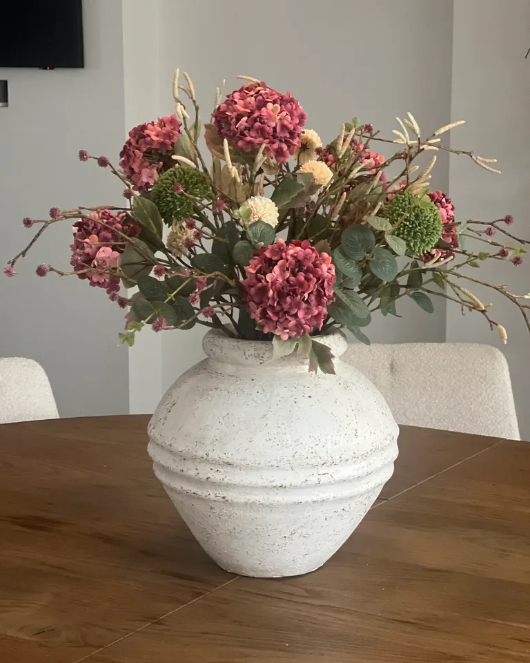 Mirelle met oudroze bloemen en groen blad in een lichte, brede vaas op een houten eettafel.