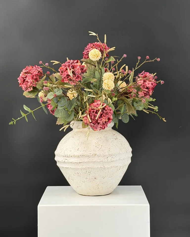 Mirelle met oudroze viburnum, crèmekleurige pompom, eucalyptus en fijne takken in een lichte vaas tegen een donkere achtergrond.