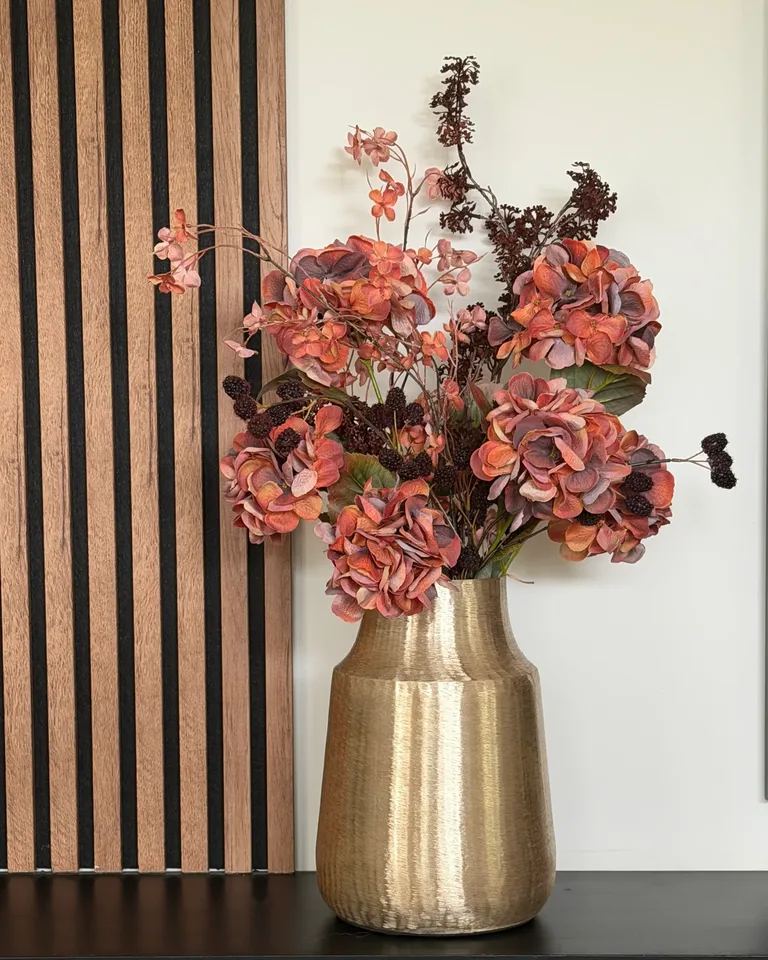 Boeket Ambra met terracotta hortensia’s en bordeauxrode bramen in een goudkleurige vaas op een zwarte wandkast, naast houten wandpanelen.