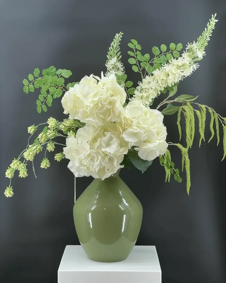 Opgaand crème-groen boeket Nerine met grote crèmekleurige hortensia’s en hangende groene takken op een witte zuil.