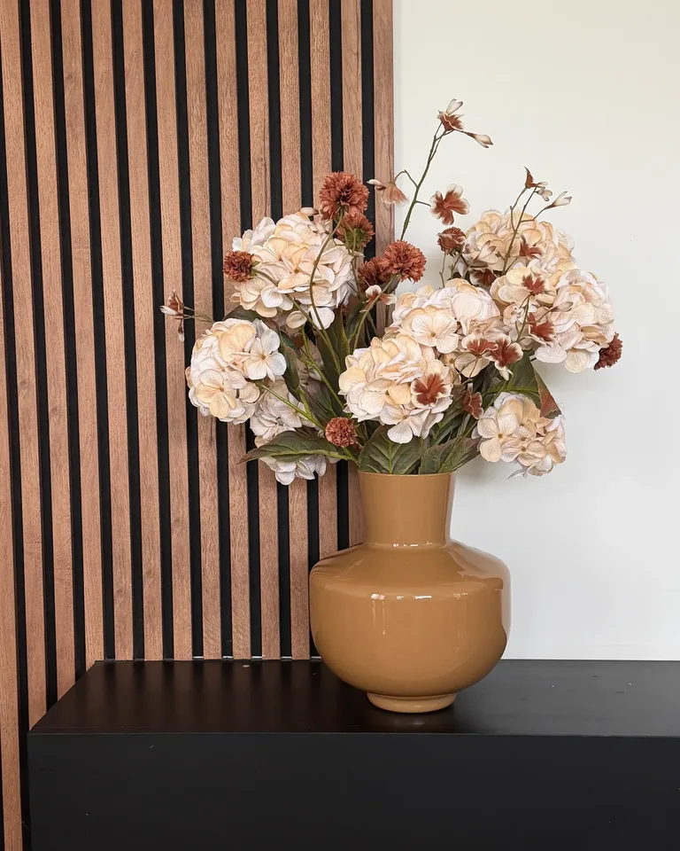 Celeste met crème hortensia en bruine bloemen in een okerkleurige vaas op een zwarte wandkast naast een houten lattenwand.