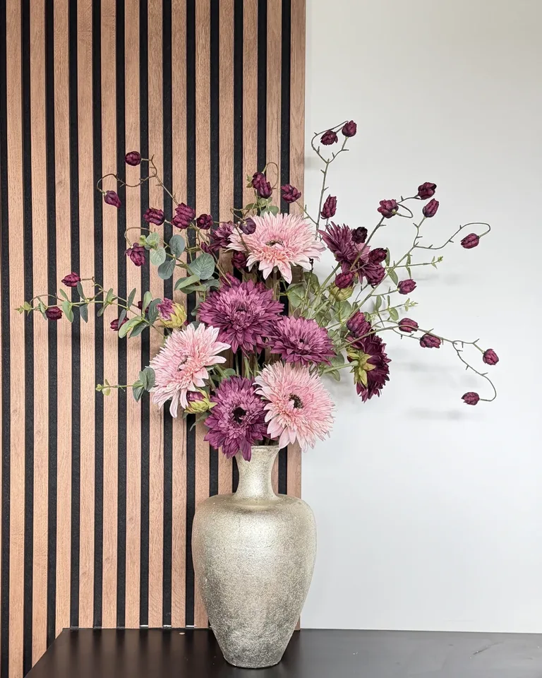 Paars-roze boeket Lunara in een goudkleurige vaas op een zwart dressoir tegen een houten lattenwand.
