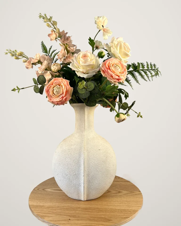 Aveline: hoog boeket met peach ranonkels, crèmekleurige rozen en groen in een lichte vaas.