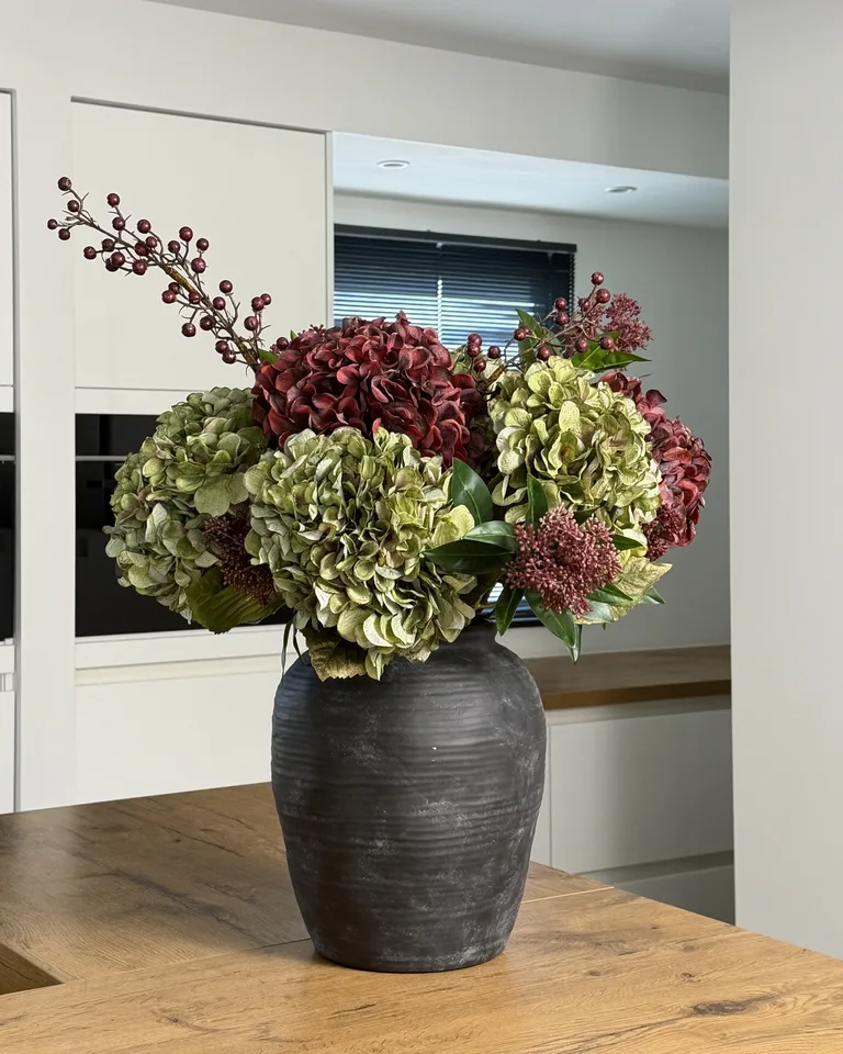 Oriane met bordeauxrode en groene hortensia's in een donkere vaas op een houten keukentafel.