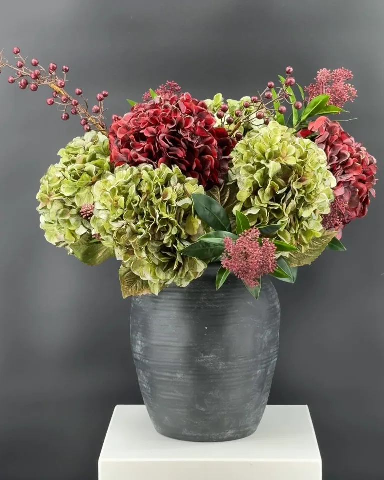 Rond boeket Oriane met bordeauxrode en groene hortensia's, skimmia en bessentakken in een donkere vaas.