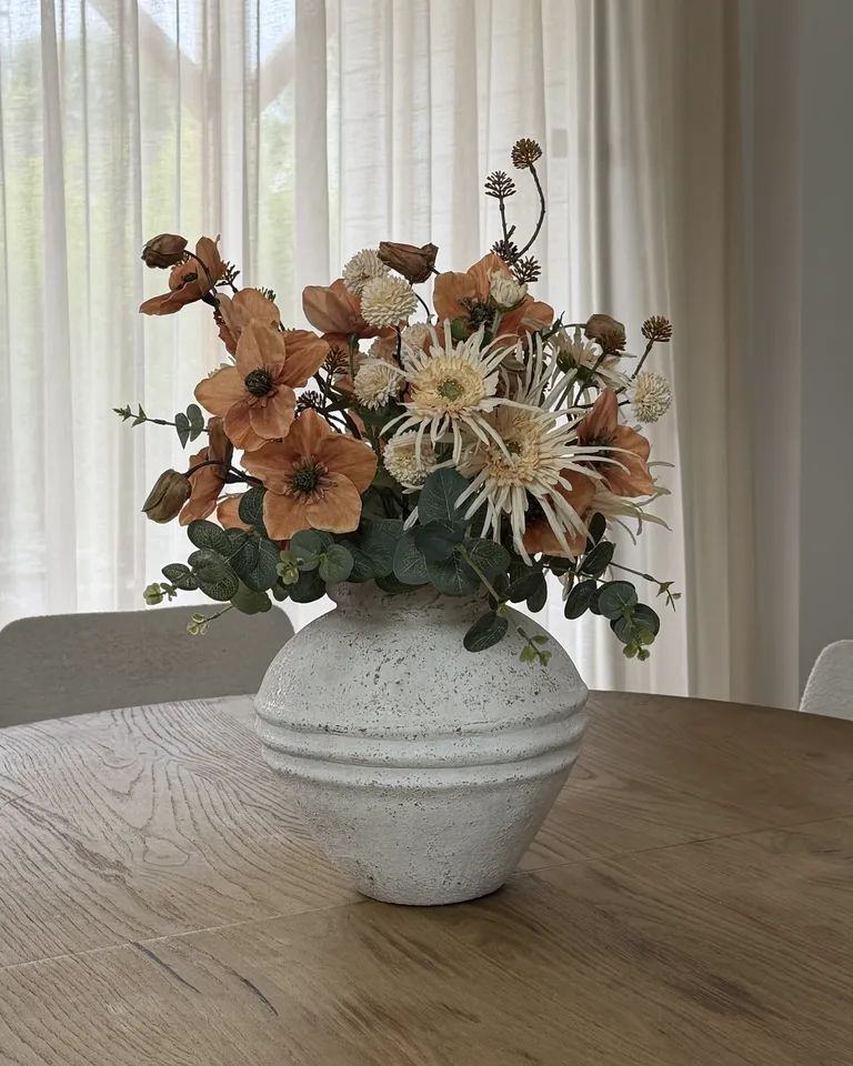 Solenne in een witte ronde vaas op een houten eettafel voor lichte gordijnen.