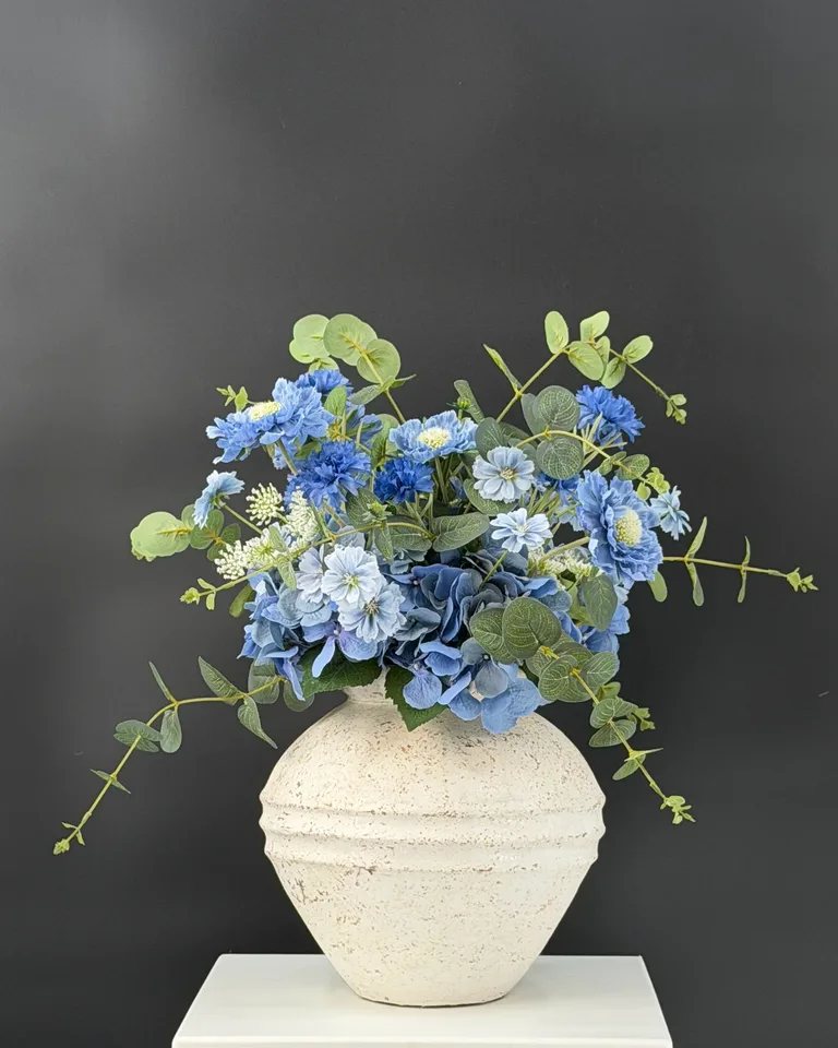 Blauw boeket met blauwe en lichtblauwe bloemen, groene takken en een witte vaas tegen een donkere studioachtergrond