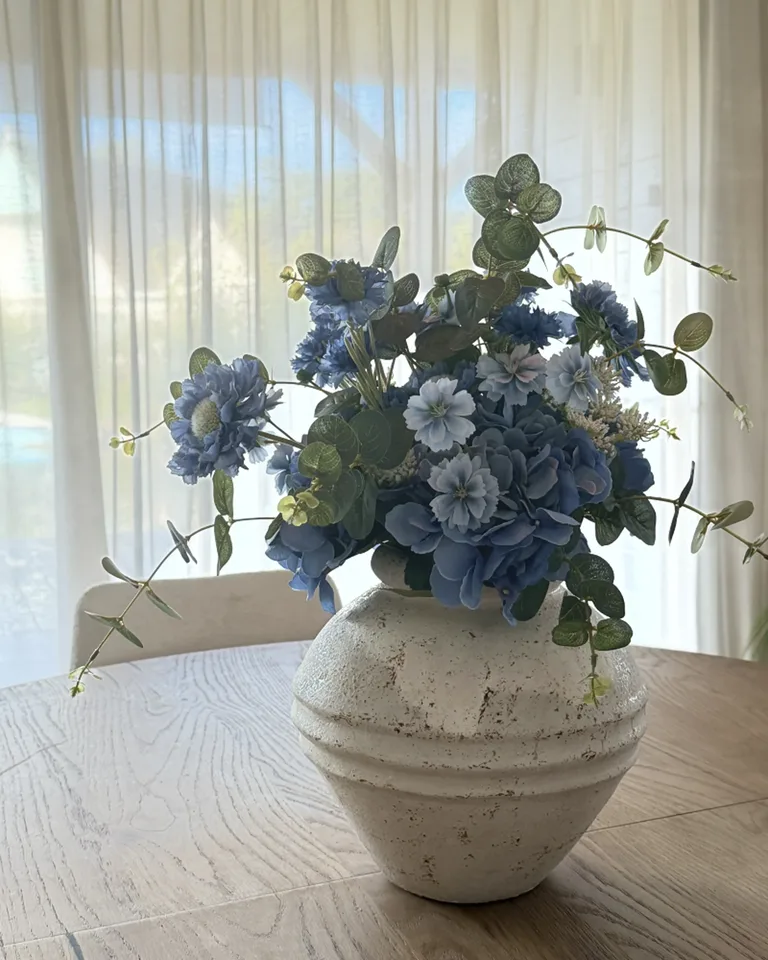 Blauw boeket met lichtblauwe en blauwe bloemen en groene takken op een eettafel voor een licht gordijn