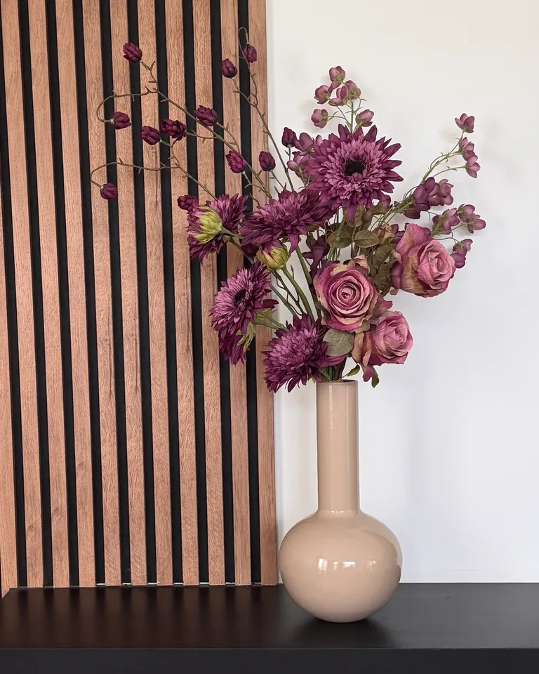 Violetta met paarse bloemen en paarsroze rozen in een beige vaas op een zwarte wandkast naast een houten lattenwand.