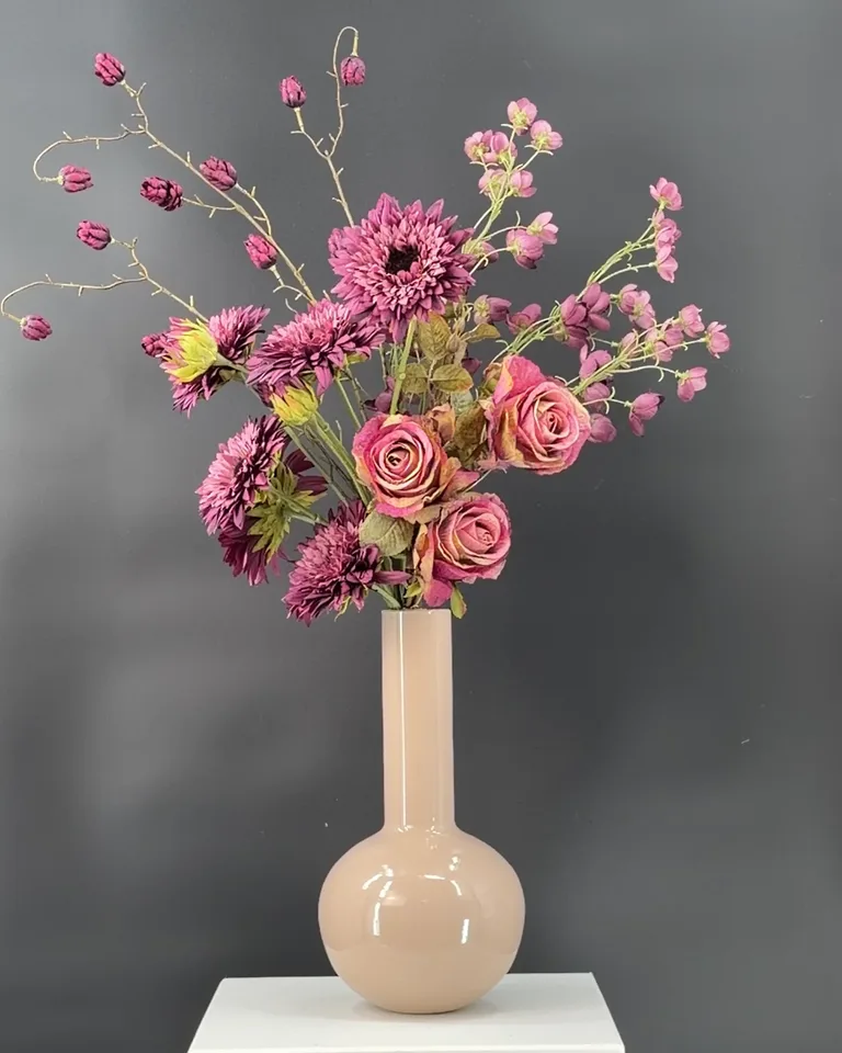 Violetta met paarse Helianthus, paarsroze rozen, buttercup en hop in een beige vaas tegen een donkere achtergrond.