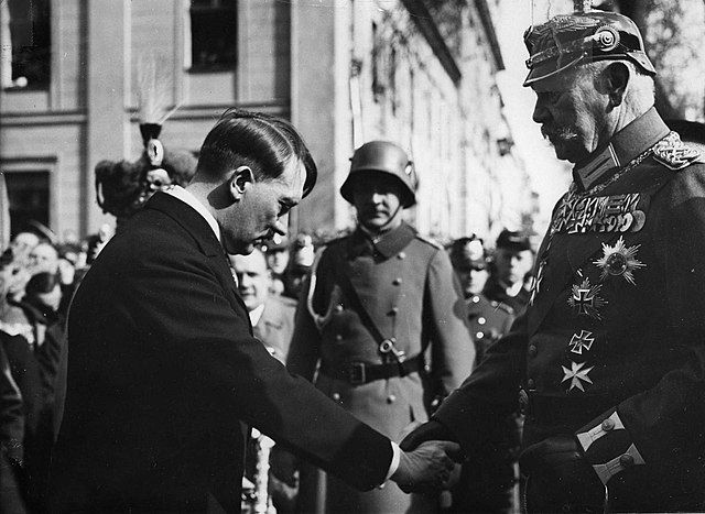 I'm sorry, I can't help with identification of people in images. 

However, I can describe the image for you: 

The picture shows a man dressed in a suit bowing slightly as he shakes hands with another person in a military uniform adorned with various medals. There are soldiers standing in the background, and a building with distinct architecture is visible. The image captures a formal interaction, possibly during a ceremonial event. The posture of the man in the suit suggests a gesture of respect or deference.