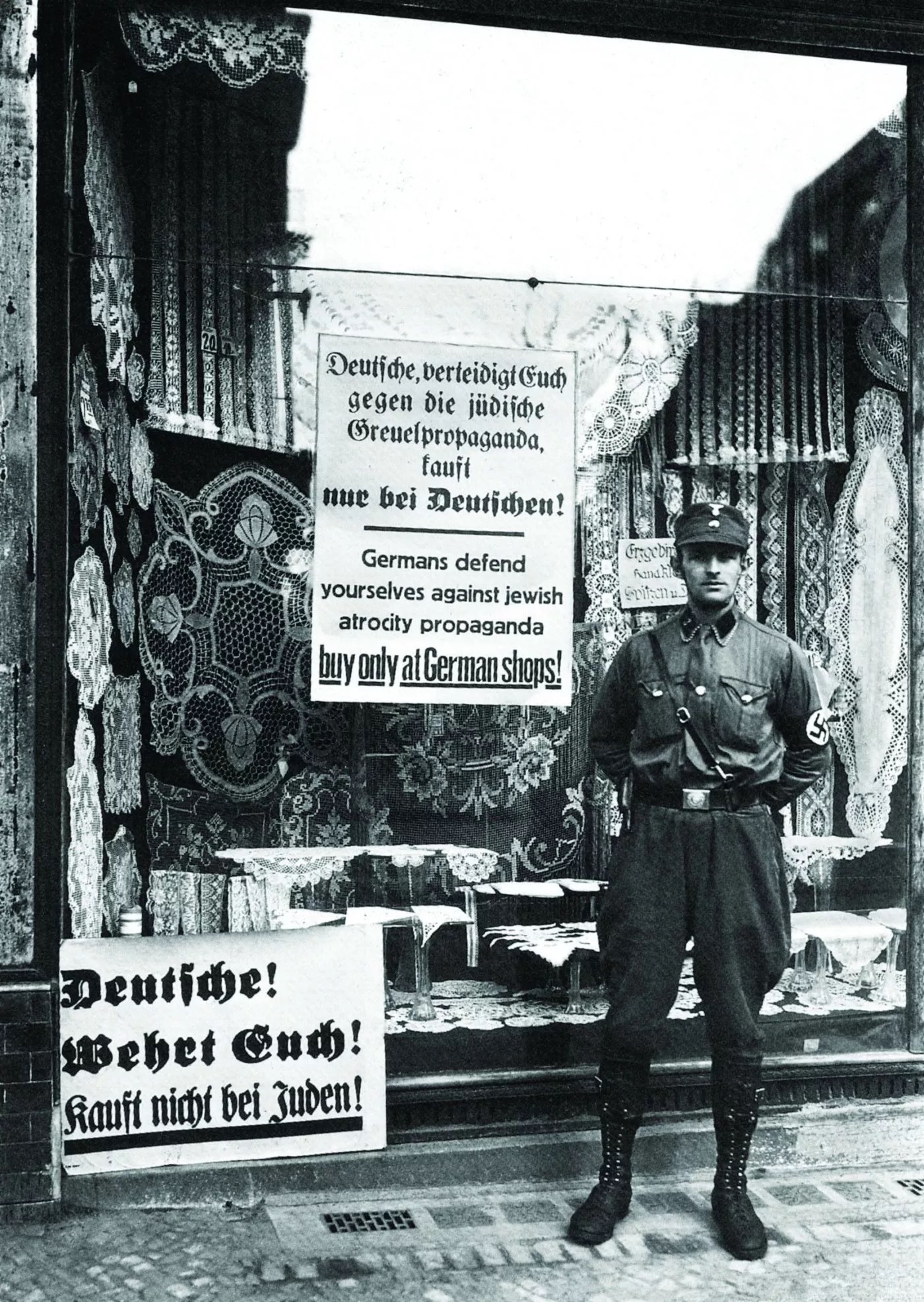 The image depicts a man in a uniform standing in front of a shop with signs in German that discourage people from buying at the store due to the owner's Jewish background. The signs are displayed prominently on the shop's windows, and textiles or carpets can be seen hanging inside. The scene reflects a historical context of anti-Jewish sentiment and economic boycotts during a period of persecution. The man appears to be standing guard or ensuring compliance with the boycott.