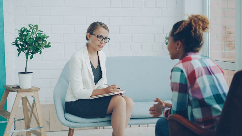 A therapist in a white blazer taking notes while listening to a client in a bright office with plants