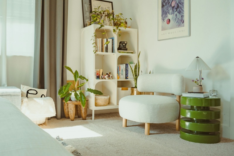 A bright, plant-filled therapy room with a rounded white bookshelf, a plush chair, and warm sunlight