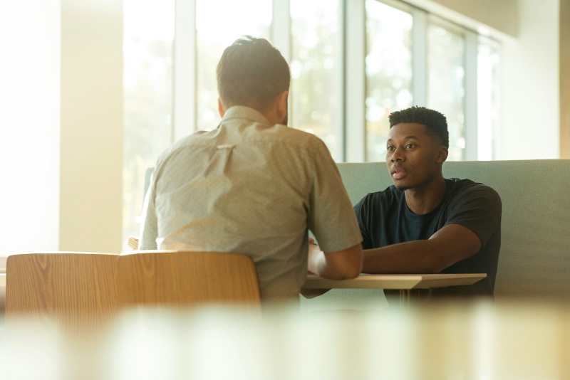 Two people in a sunlit, open conversation across a table — one listening intently, one speaking