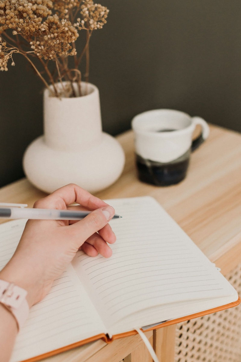 A hand holding a pen over an open journal on a wooden surface, with a mug and dried flowers nearby