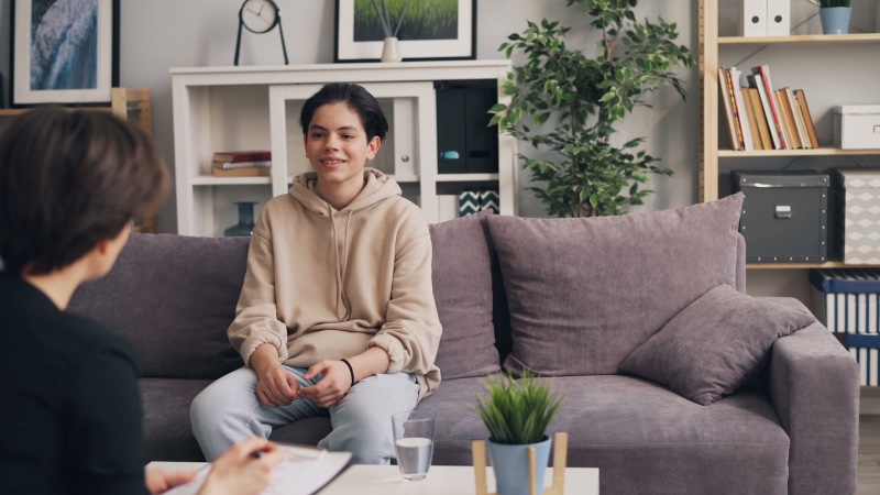 A teenager in a hoodie sitting on a therapy office couch, speaking with a clinician