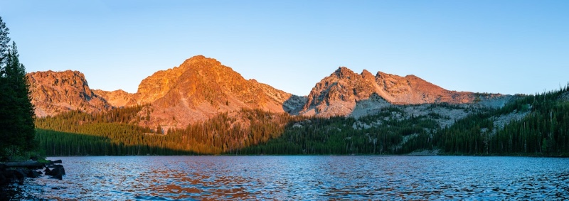 A calm alpine lake reflecting alpenglow on rugged mountain peaks and pine forests