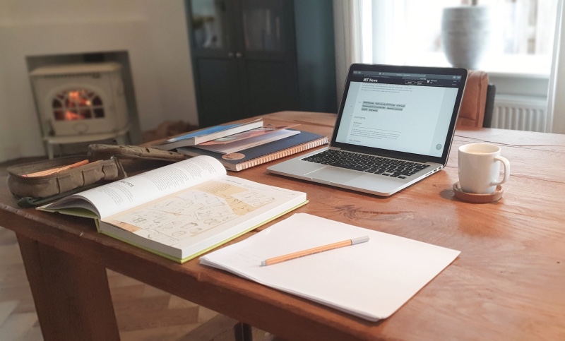 An open laptop, books, a notepad, and a mug on a wooden table near a glowing fireplace