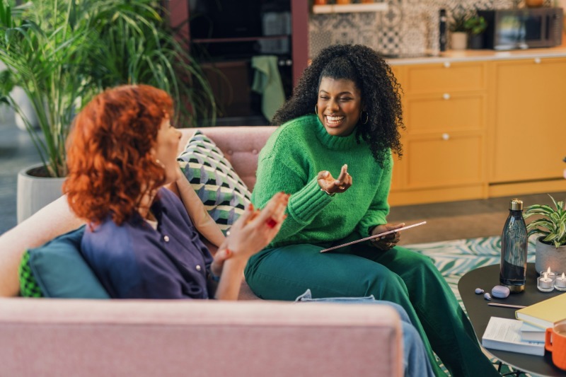 Two women talking warmly on a sofa in a bright, plant-filled room