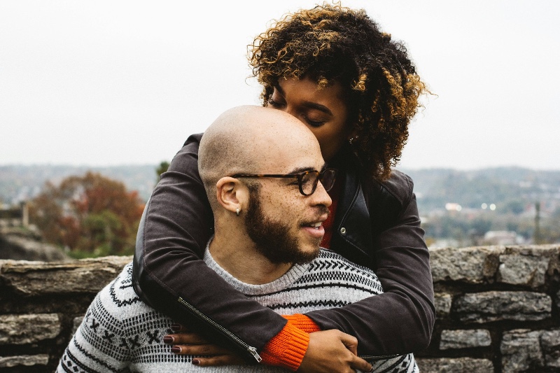 A couple in a close, tender embrace outdoors — one resting their head against the other's