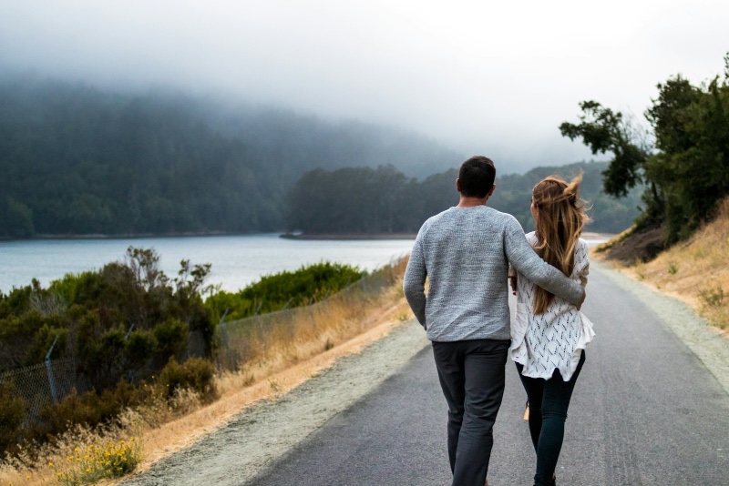 A couple walking arm-in-arm along a misty road beside a foggy lake