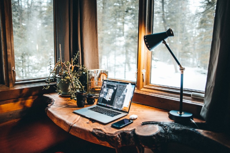A computer sits at a desk in a rustic cabin in the woods with snow outside