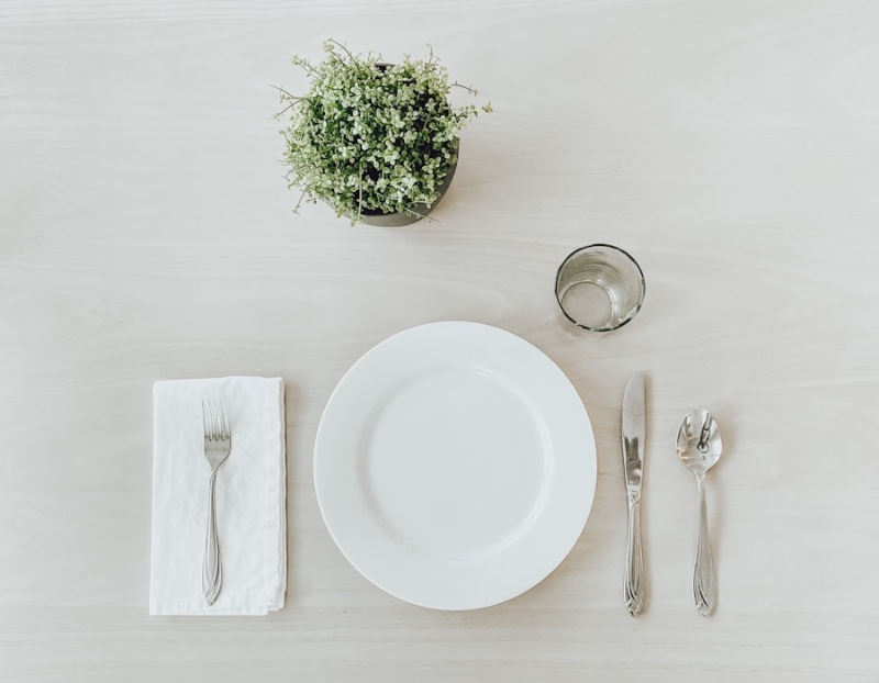 A minimal place setting from above — white plate, silverware, water glass, and a small green plant