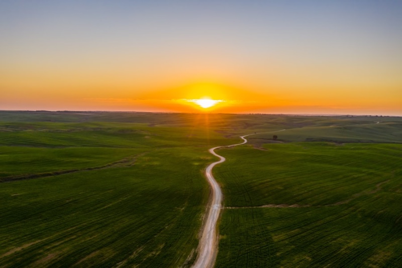 An aerial view of a winding country road through vast green rolling fields toward a golden sunset