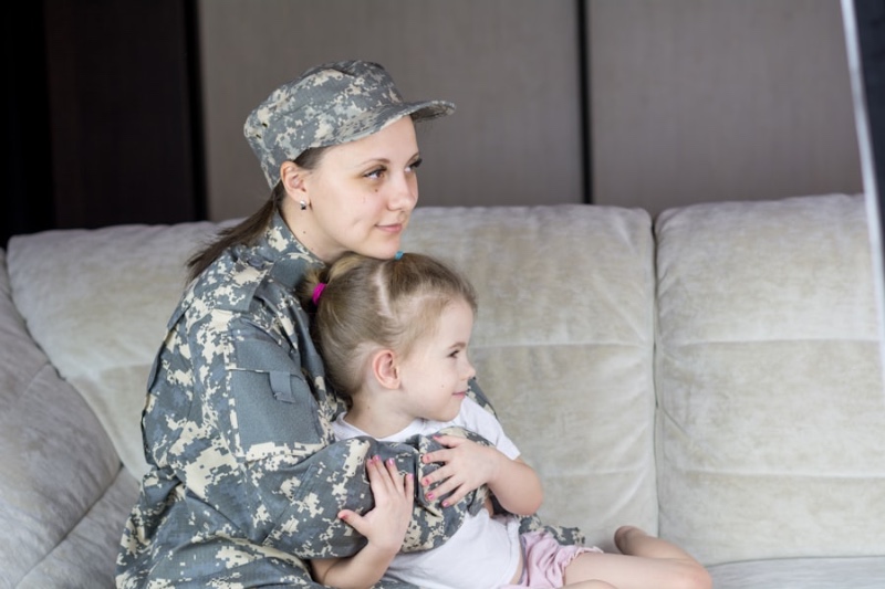 A woman in military camouflage holding a young child on a couch, both gazing gently to the side
