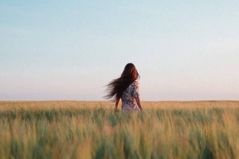 A woman seen from behind in a floral dress, standing in a golden grass field with her hair blowing in the breeze