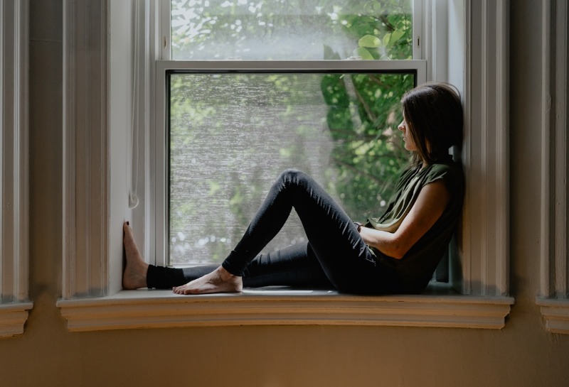 A person sitting alone on a wide window ledge, looking out at the trees
