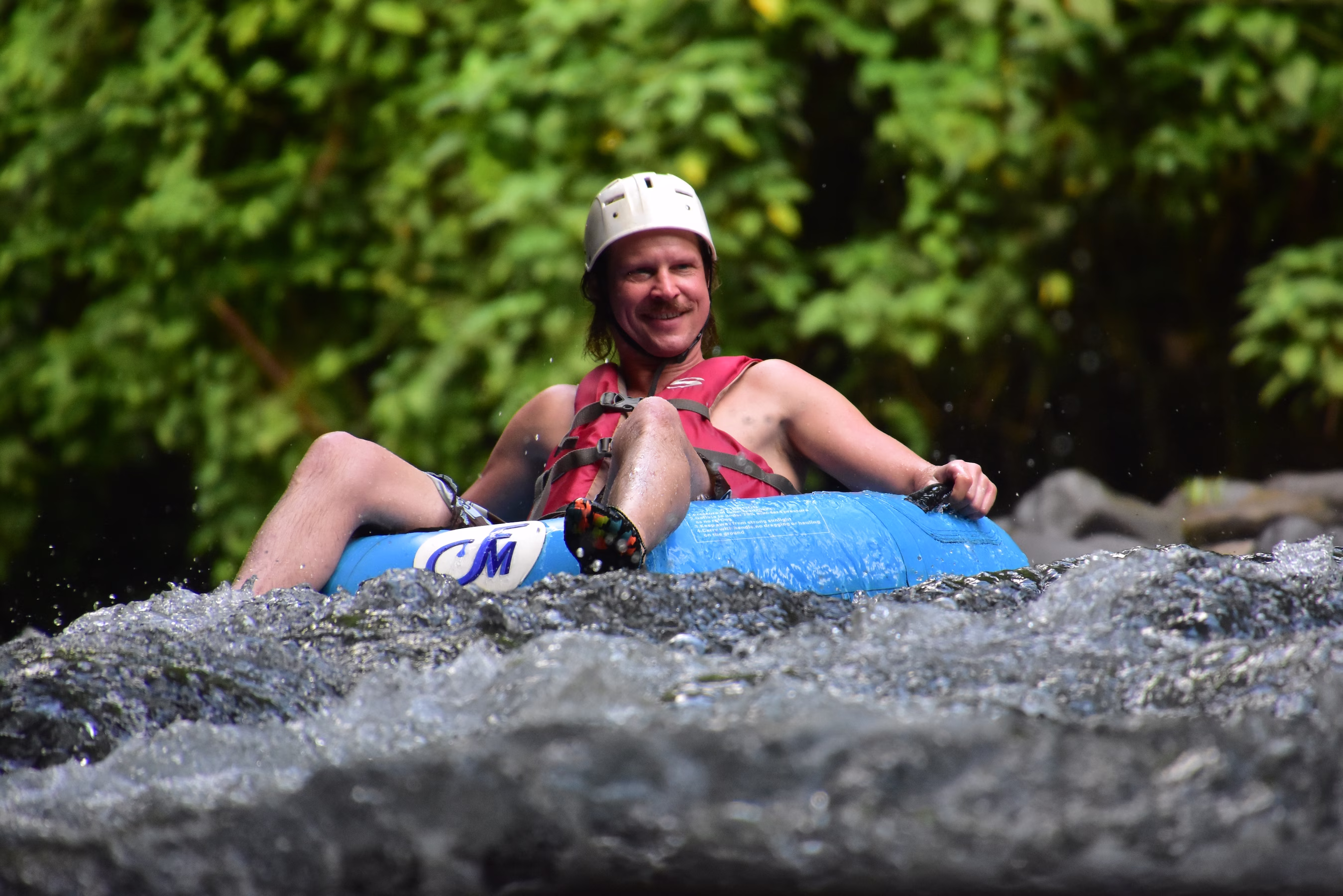 Man relaxing on a blue tube in the river, smiling at the camera