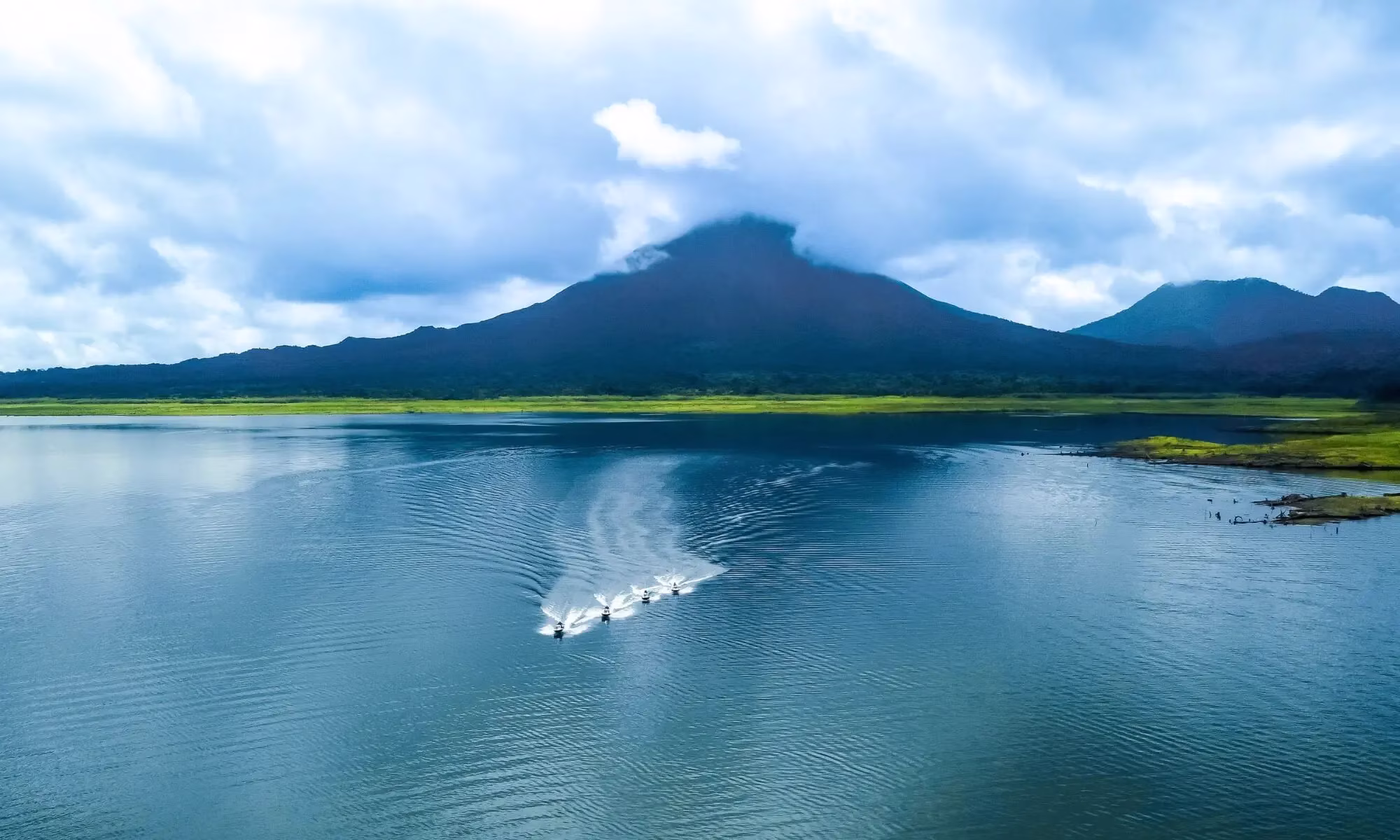 Drone view of four people riding jet skis on Lake Arenal with Arenal Volcano in the background, its summit covered by clouds, Costa Rica